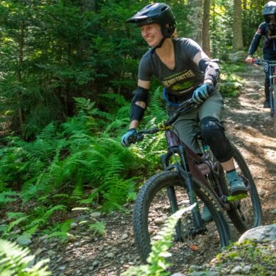 A women mountain biking with a guy behind her. Lots of ferns.