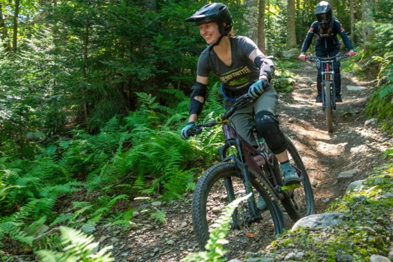 A women mountain biking with a guy behind her. Lots of ferns.