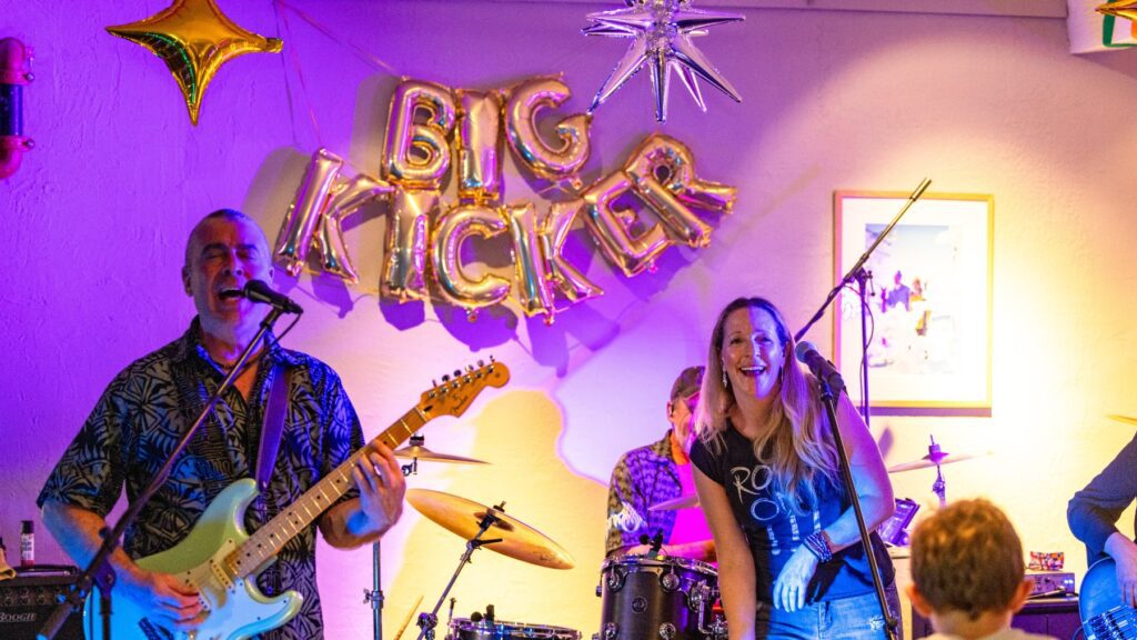 A band playing in front of blown up bubble letters that says The Big Kicker 
